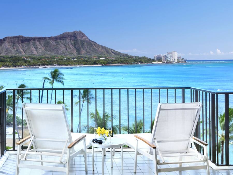 Ein Balkon mit zwei weißen Liegestühlen, einem kleinen Tisch und Blick auf Strand und Berge.