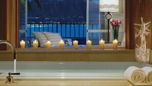 A bathtub with candles, a window with a view of a mountain and a beach at night, a lamp, and a vase with flowers. - Halekulani (Honolulu)