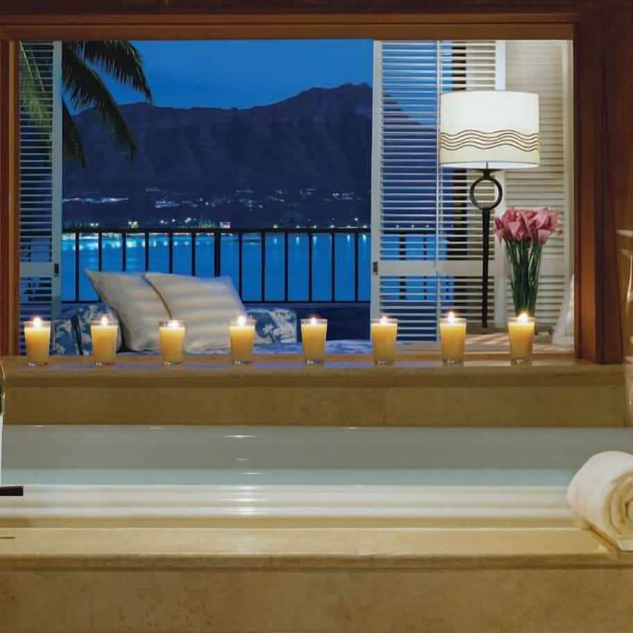 A bathtub with candles, a window with a view of a mountain and a beach at night, a lamp, and a vase with flowers.