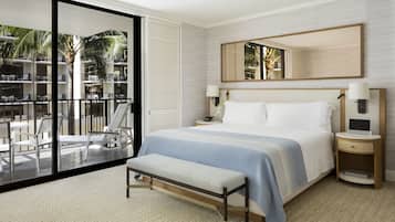 A modern hotel room with a large bed, a bench, a nightstand, and a large window offering a view of the outdoor seating area.