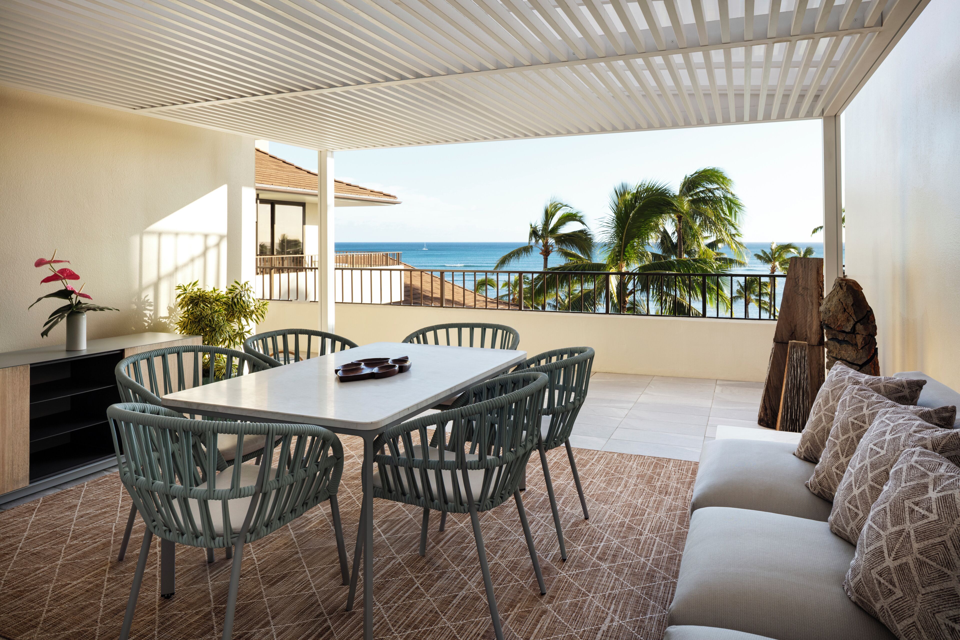 A balcony with a view of the ocean, a dining area with chairs and a table, and a sofa with patterned cushions.
