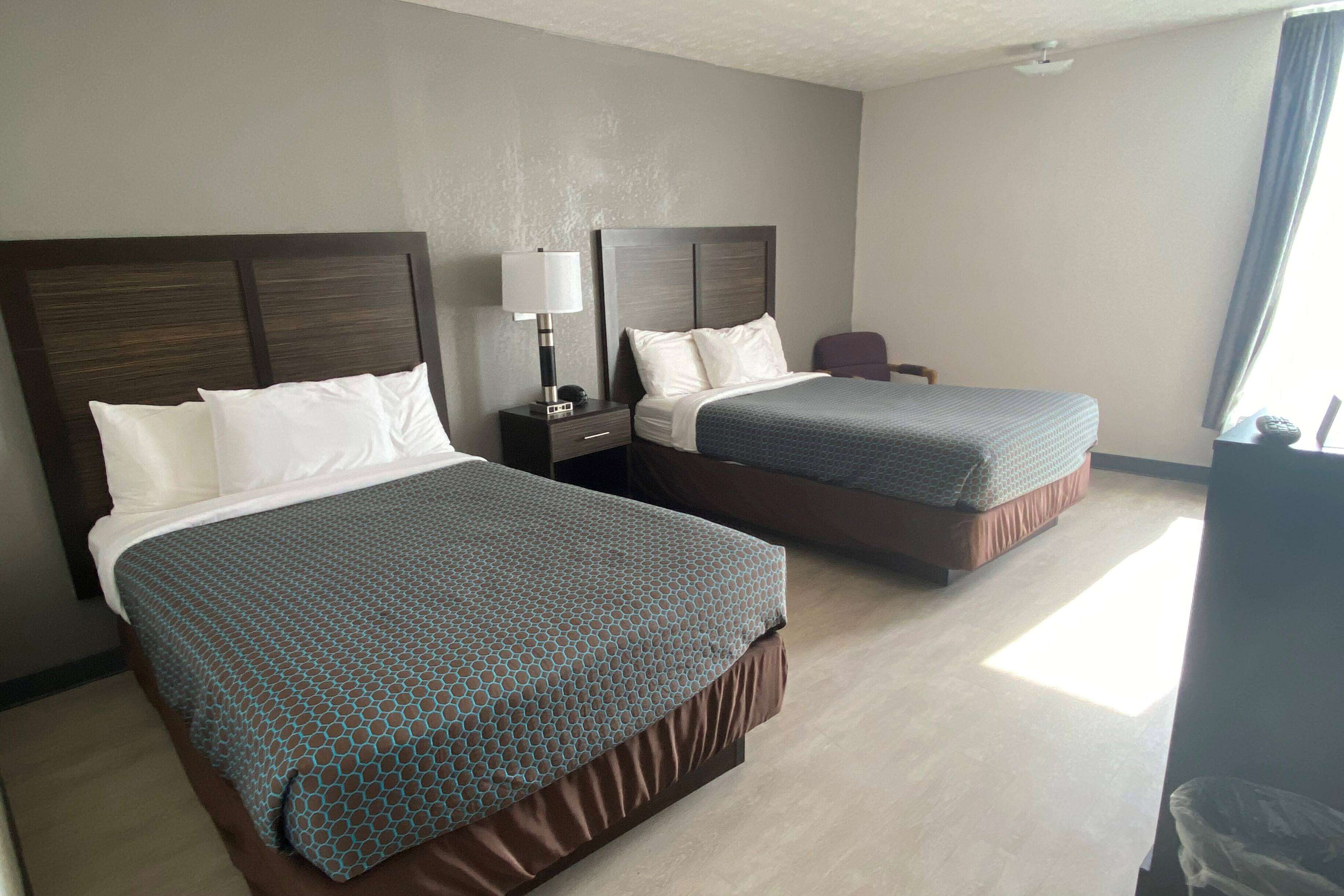 A hotel room with two beds, a nightstand, a lamp, and a chair.