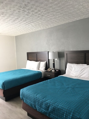 A hotel room with two beds, each with a blue patterned bedspread, a wooden headboard, a bedside table with a lamp, and a telephone.