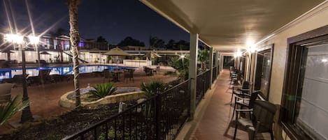 Una vista notturna di un balcone d'hotel con piscina, posti a sedere all'aperto e vista sugli edifici circostanti.