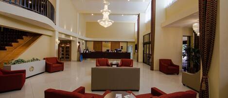 A spacious lobby with a reception desk, red seating, a chandelier, and a staircase.