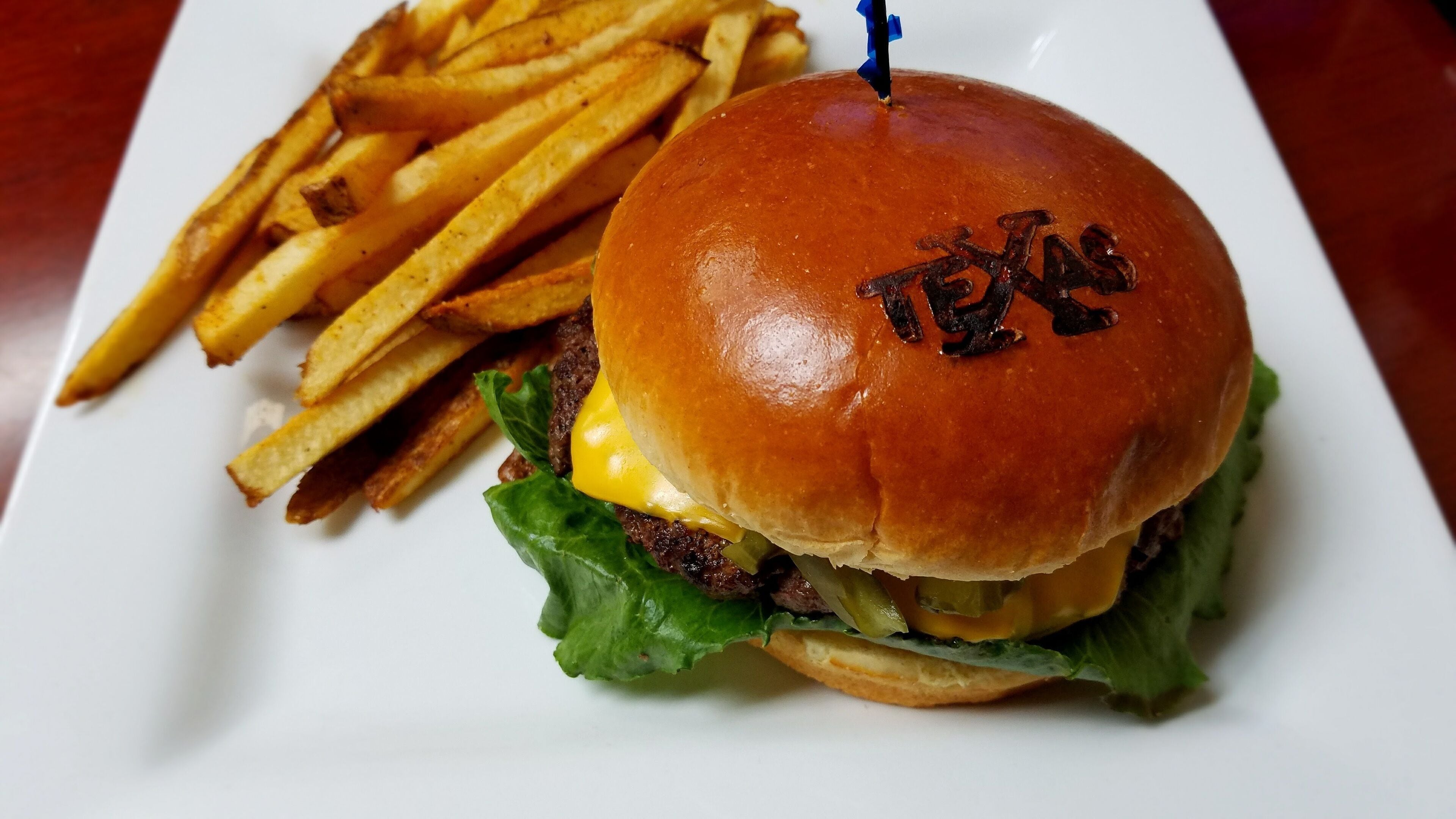 Ein Burger mit einem Brötchen, das 'TEXAS' beschriftet ist, und eine Portion Pommes auf einem weißen Teller.