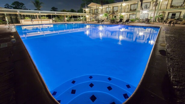 A swimming pool with a clear blue water surface, surrounded by a covered area with seating and a building in the background.