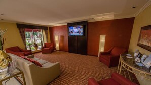 A hotel lobby with a large flat-screen TV mounted on the wall, multiple red upholstered chairs, a beige sofa, a glass table with a magazine rack, and a patterned carpet.