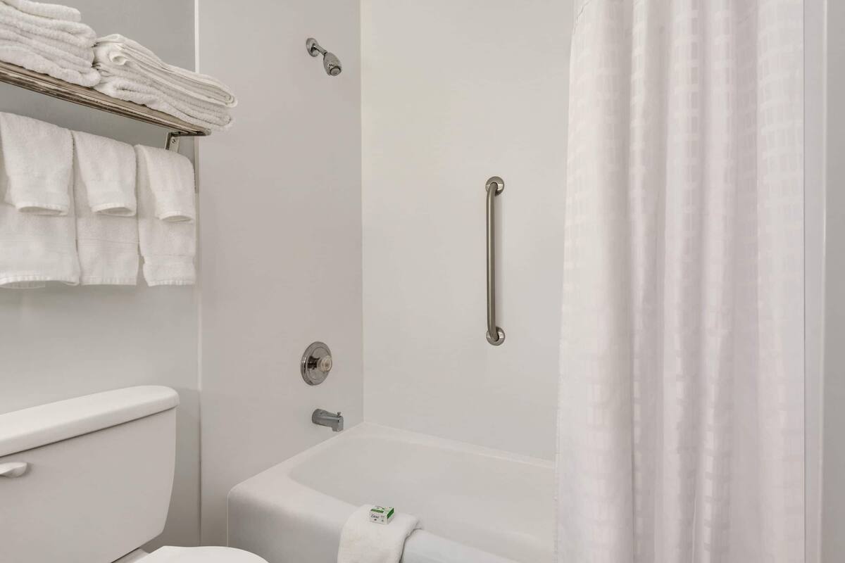Un baño con inodoro, bañera, ducha con barra de apoyo y toallero con toallas dobladas.