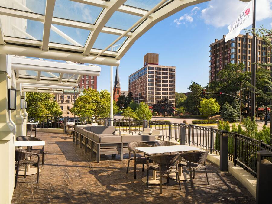 An outdoor seating area with tables and chairs, a glass roof structure, and a view of the cityscape including buildings and trees.