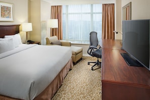 Ein Hotelzimmer mit einem großen Bett, einem Schreibtisch mit Fernseher, einem Sessel und einem Fenster mit Vorhängen.