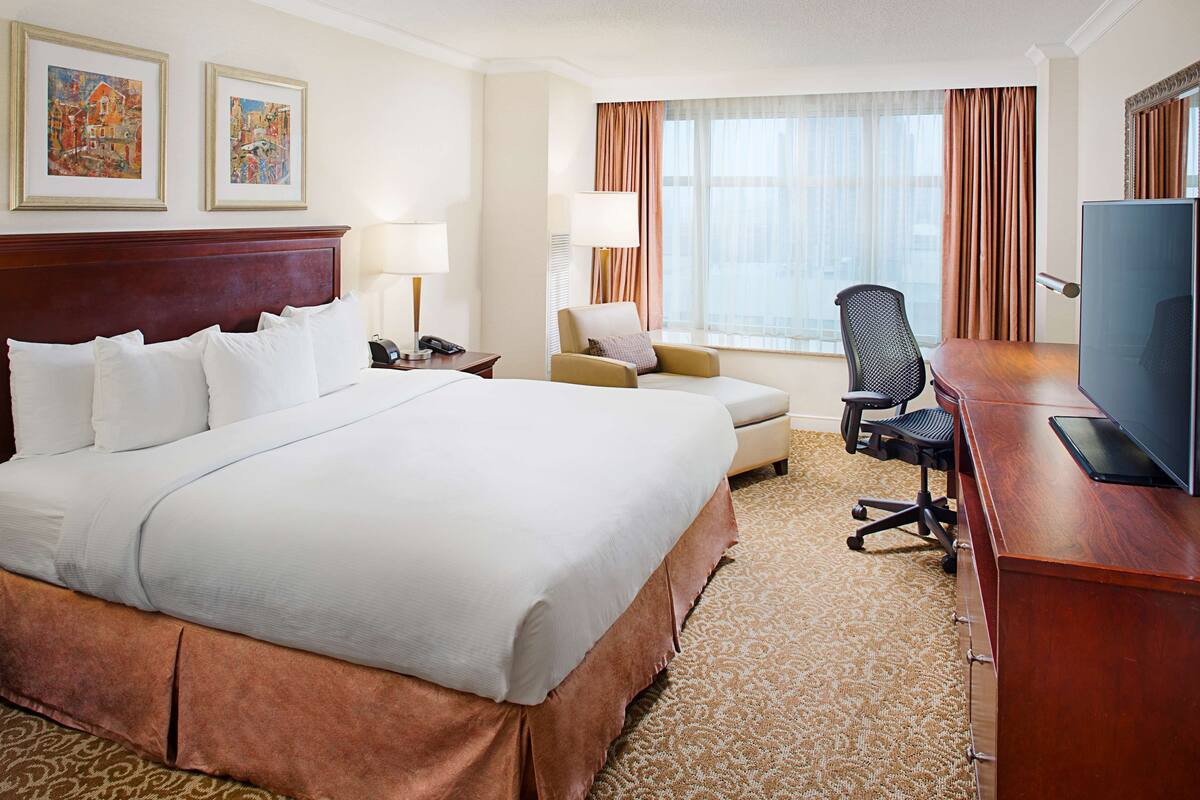 A hotel room with a large bed, a desk with a television, a chair, and a window with curtains.