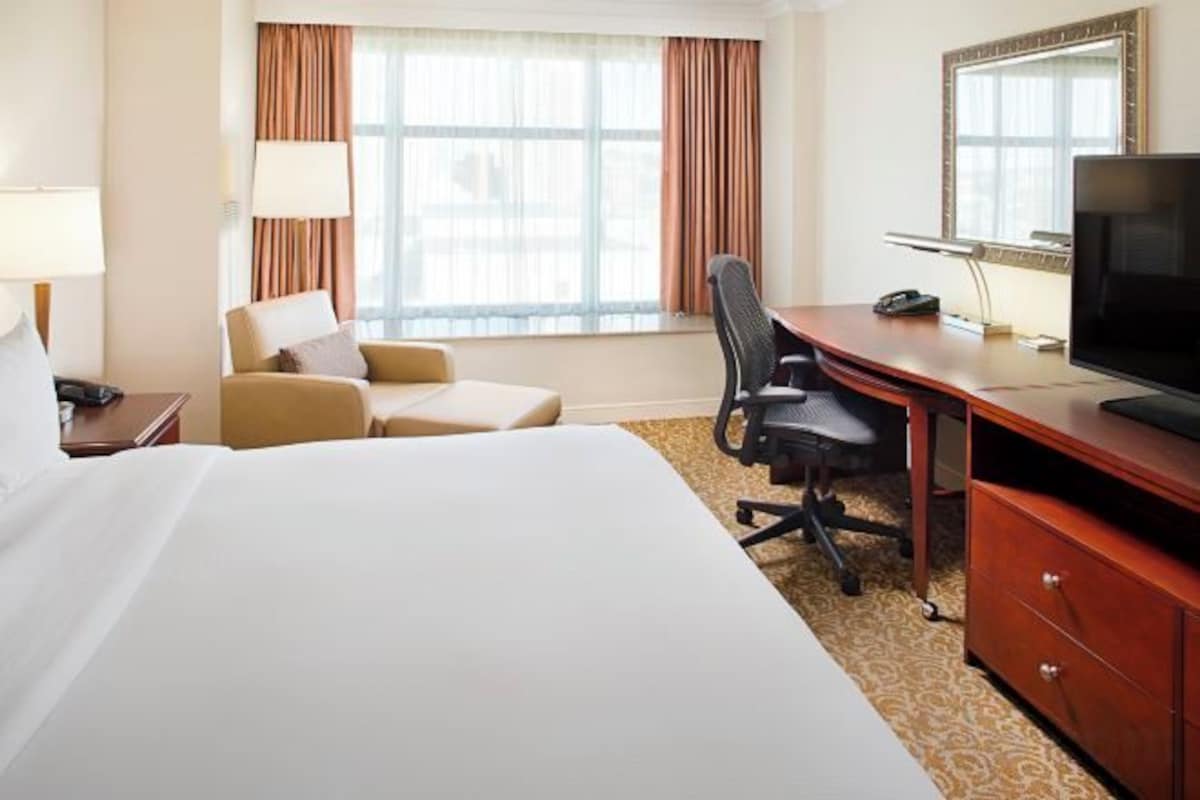 Habitación de hotel con una cama grande, un escritorio con silla, un televisor y una ventana con cortinas.
