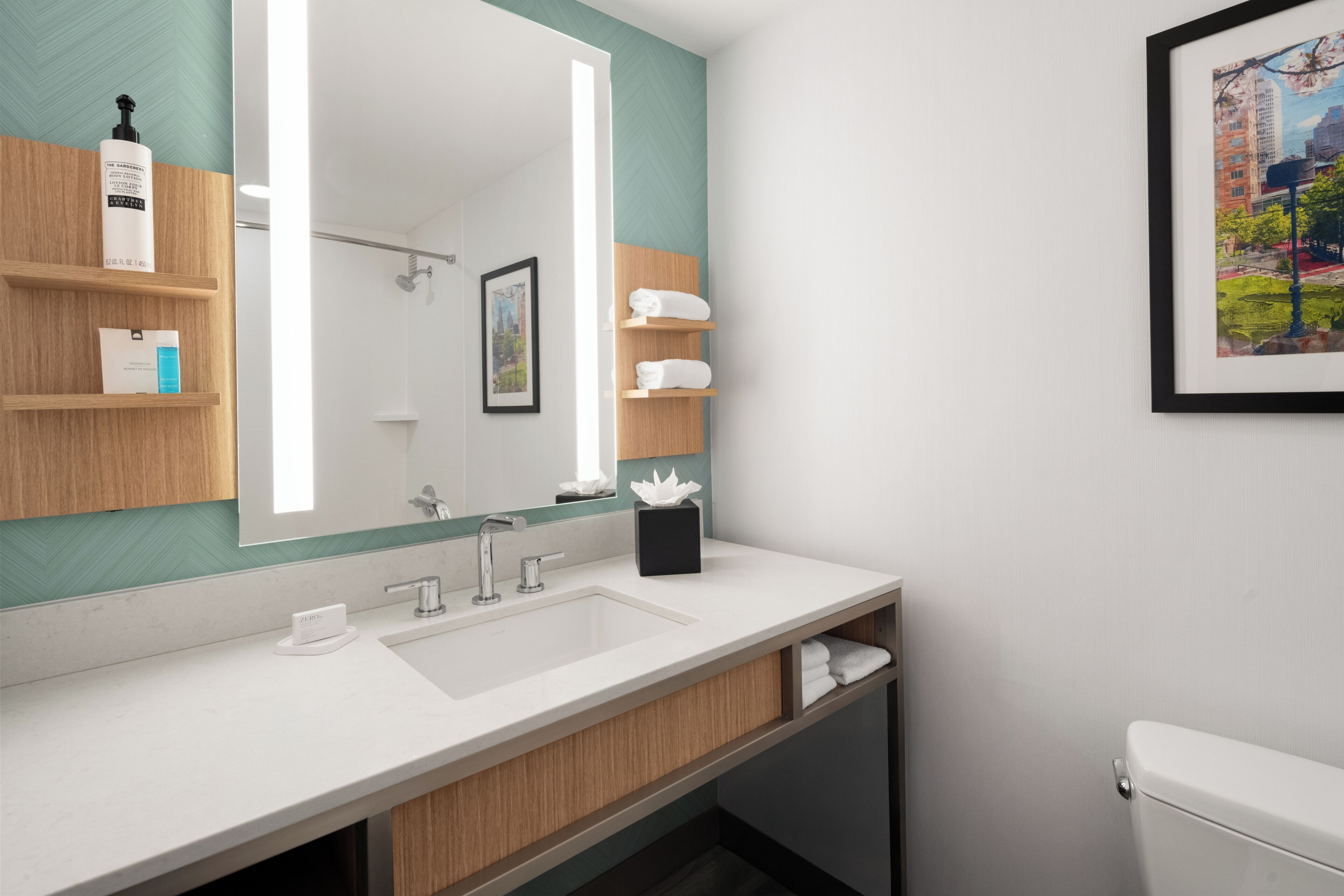 Un baño moderno con un espejo grande, un lavamanos blanco y un inodoro. Hay repisas de madera con artículos de tocador y un cuadro en la pared.