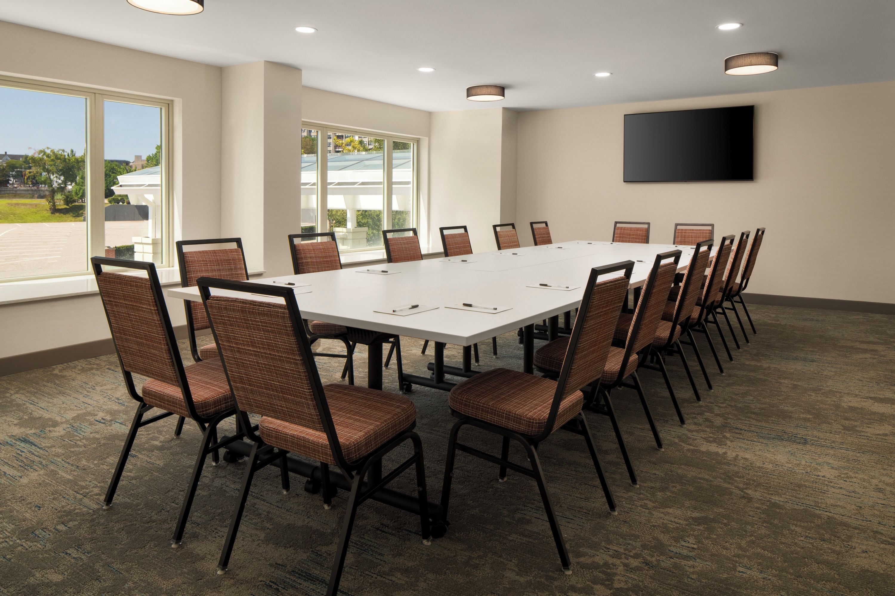 A conference room with a long table and chairs, large windows, and a flat-screen TV on the wall.
