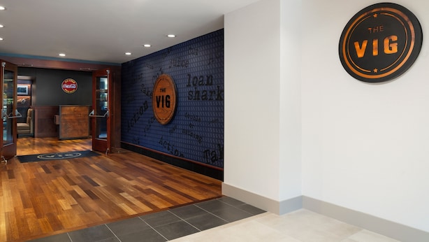 Un área de recepción con piso de madera, pared con diseño azul y un letrero circular que dice 'THE VIG'.
