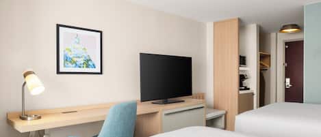 Habitación de hotel con cama, escritorio con computadora, televisor, lámpara y un cuadro en la pared.