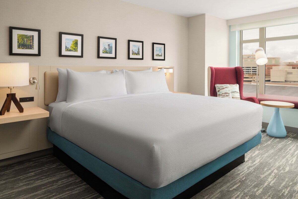 A hotel room with a large bed, a desk with a lamp, a chair, and a window with a view of buildings.