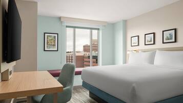 A hotel room with a bed, a desk, a chair, a TV, and a window with a city view.