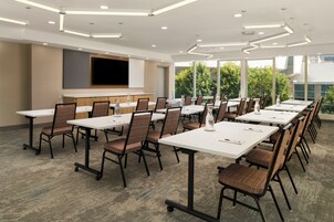 Un salón de conferencias con varias mesas y sillas, un televisor de pantalla plana montado en la pared y grandes ventanales con vistas a zonas verdes.