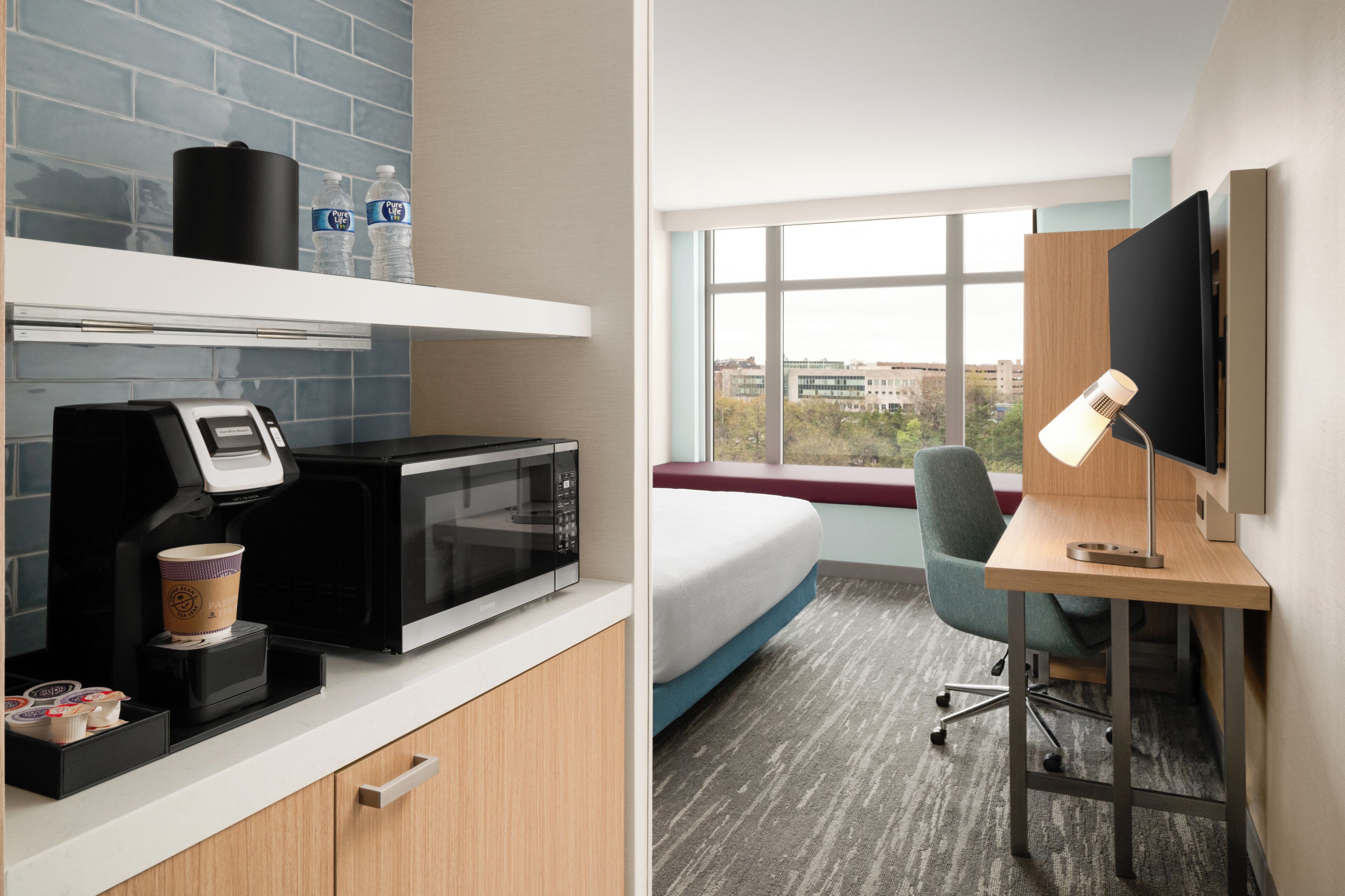 A hotel room with a kitchenette, a bed, a desk with a computer and lamp, and a window with a city view.