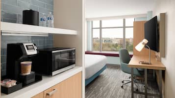 A hotel room with a kitchenette, a bed, a desk with a computer and lamp, and a window with a city view.