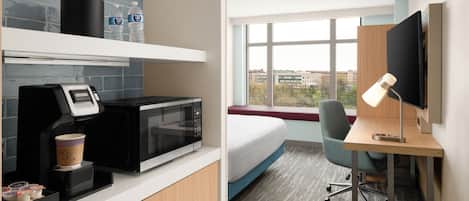Quarto de hotel com cozinha compacta, uma cama, escrivaninha com computador e luminária, e janela com vista para a cidade.
