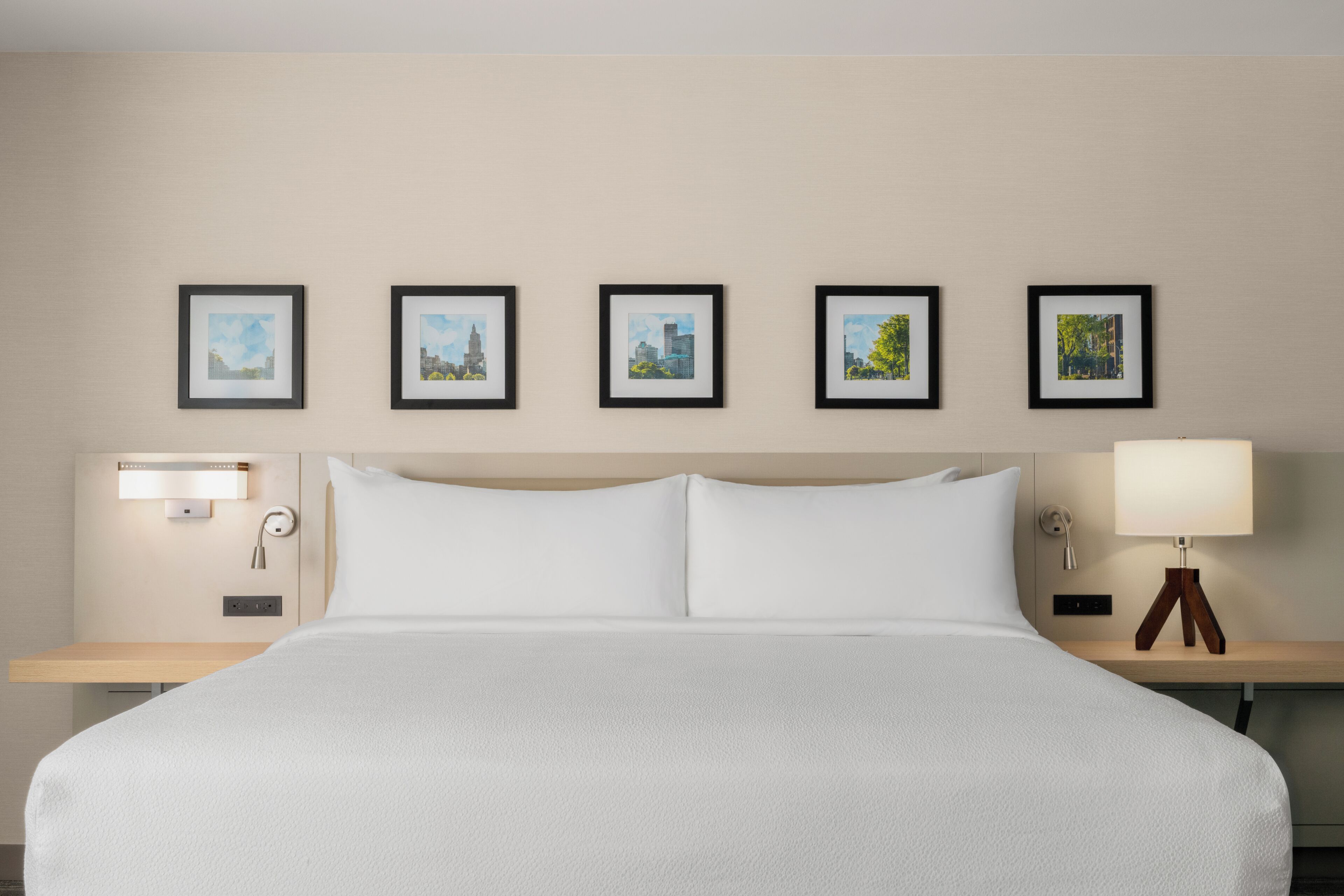 Une chambre d’hôtel avec un grand lit, une table de chevet avec une lampe et cinq tableaux encadrés accrochés au mur.