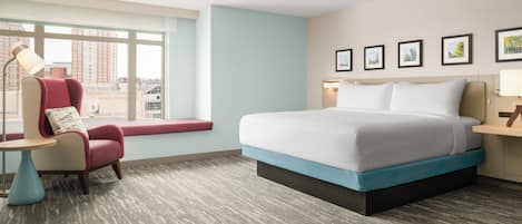 A hotel room with a bed, a chair, a desk, a window, and framed pictures on the wall.