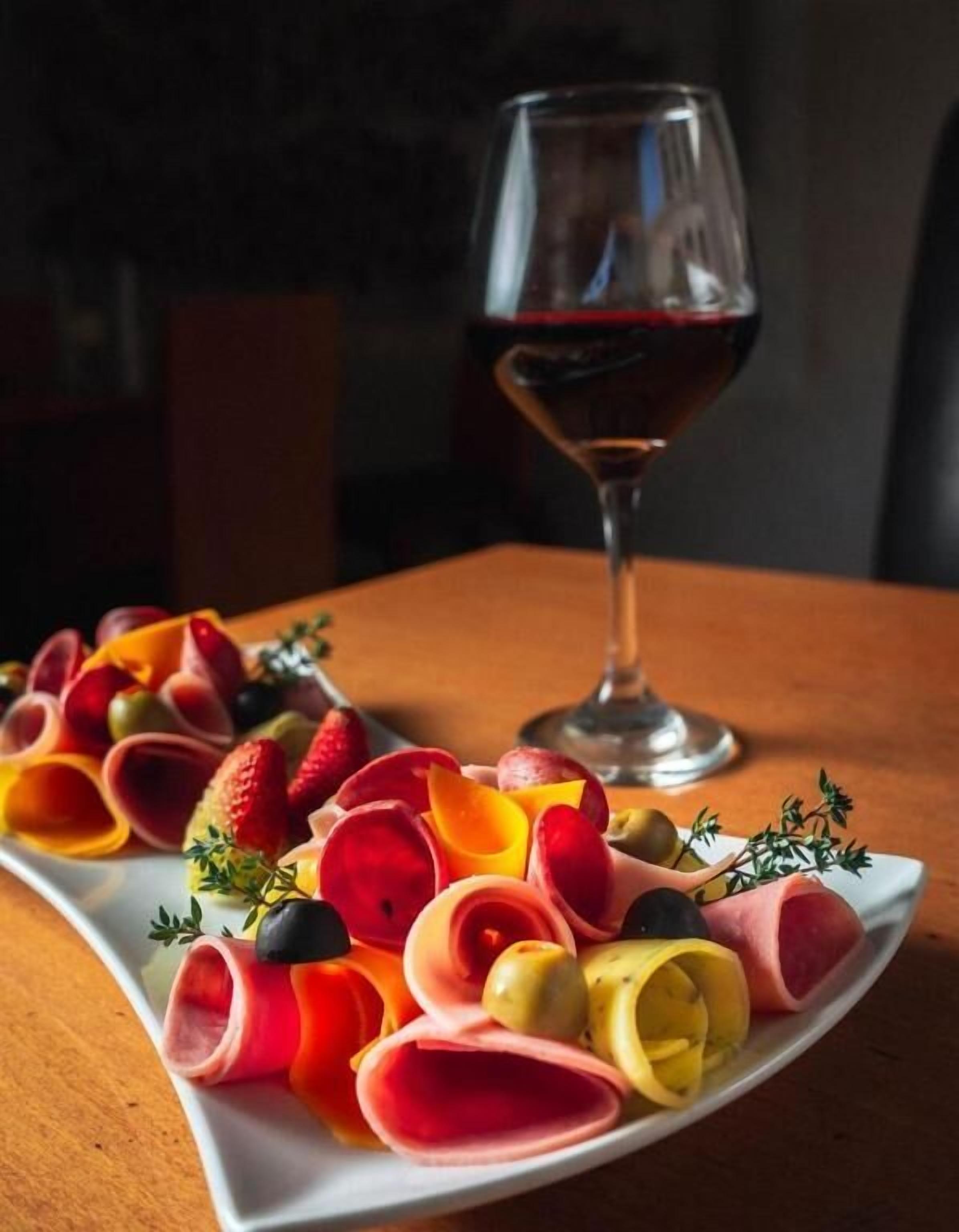 En tallerken med udvalgte spegepølser og frugter, et glas rødvin og en kvist timian på et bord.