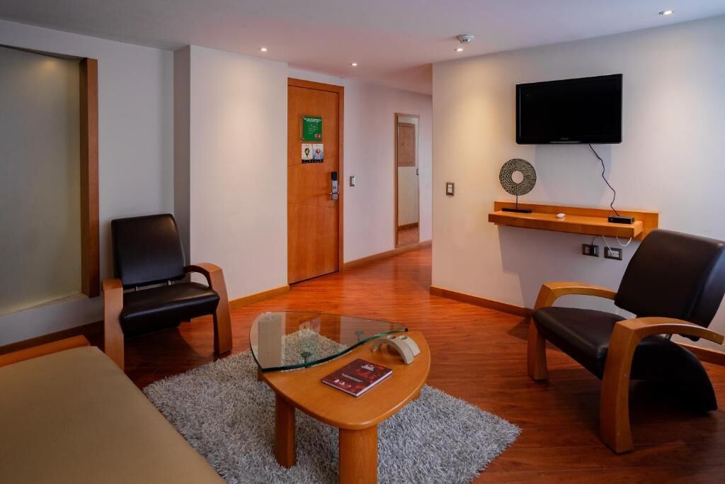Une chambre d’hôtel avec un plancher en bois, une table en verre, deux chaises noires, un téléviseur fixé au mur et une porte avec une enseigne verte.