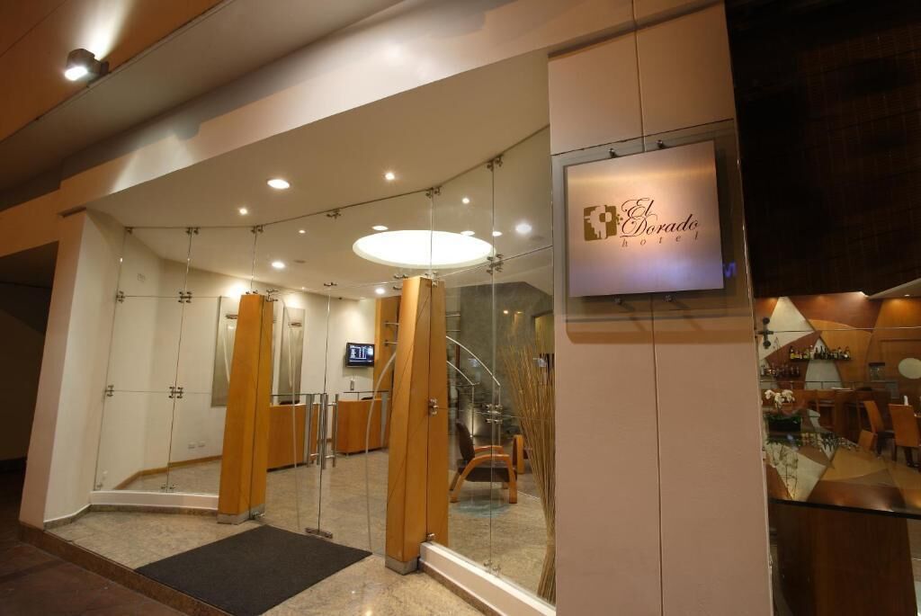 A hotel entrance with a reception desk, glass doors, and a staircase leading to the interior.