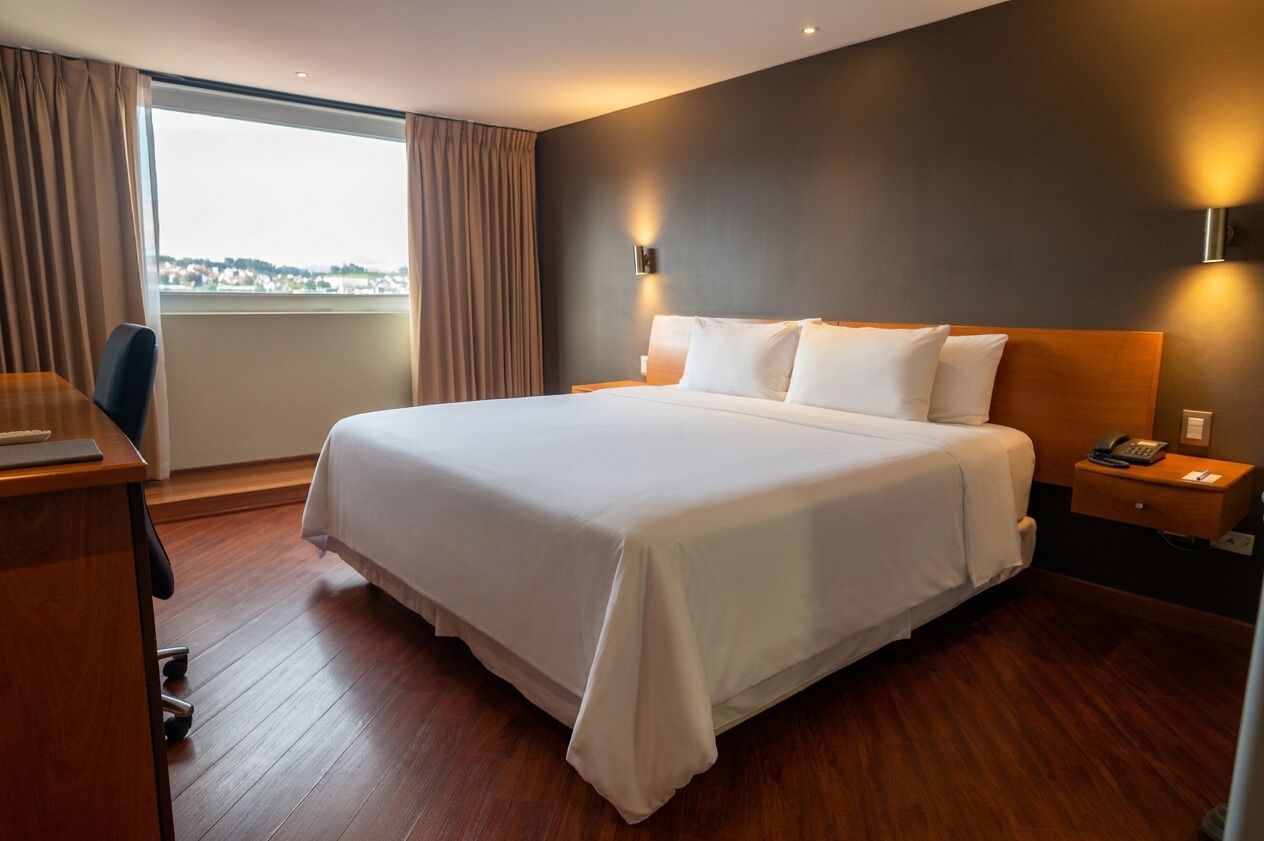 Habitación de hotel con una cama grande, un escritorio, una silla, una mesita de noche con teléfono y vistas a la ciudad desde la ventana.