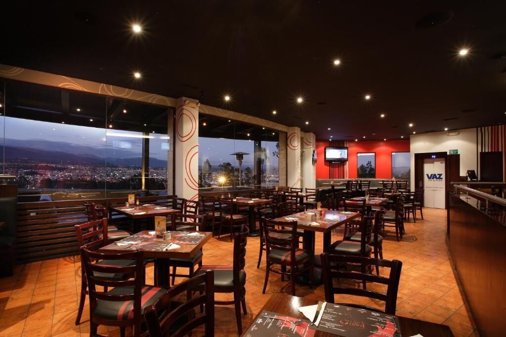 Un restaurante con varias mesas y sillas, amplios ventanales que ofrecen vistas a la ciudad, y un letrero VAZ en la pared.