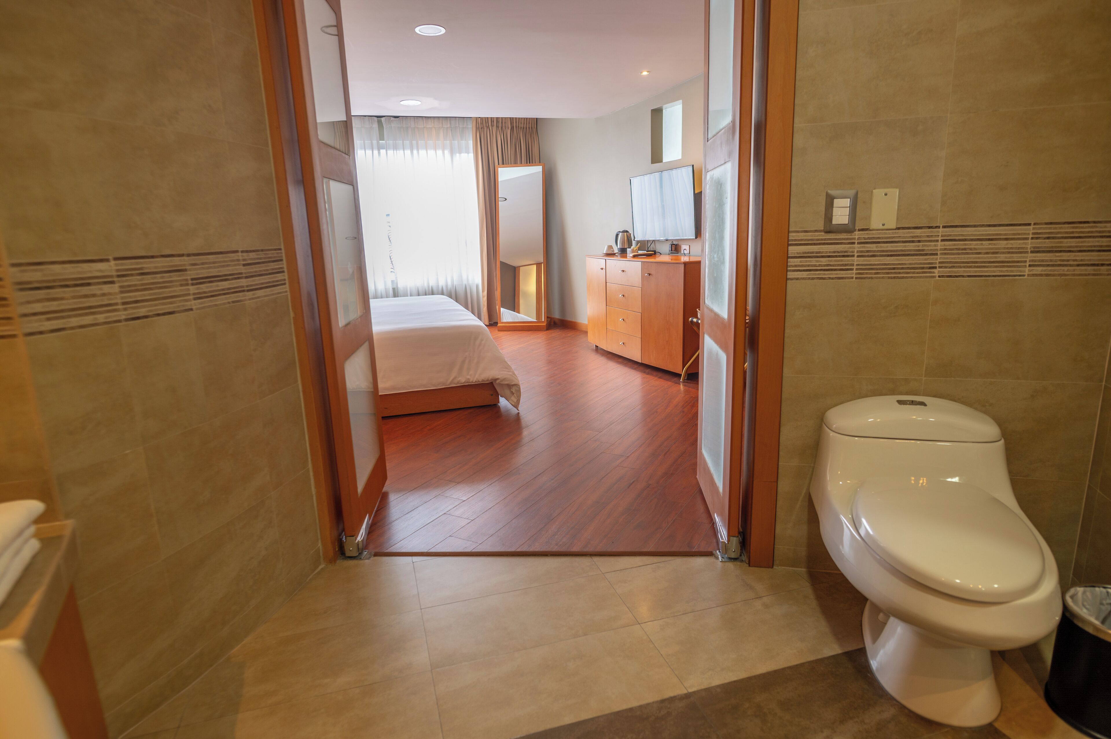 A hotel room with a bed, a bathroom with a toilet, a mirror, and a television.