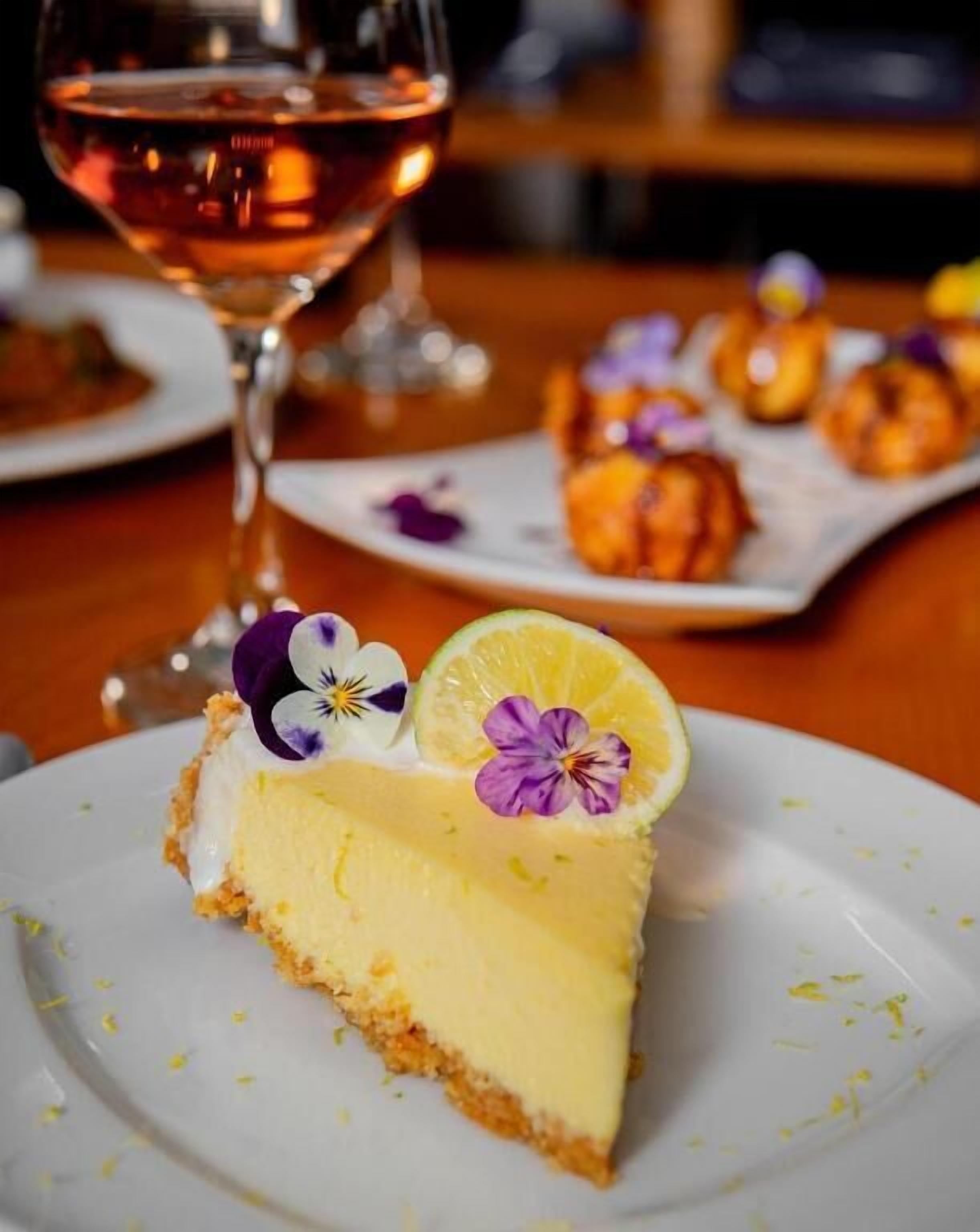 A slice of cheesecake on a plate with a slice of lemon and edible flowers on top, accompanied by a glass of rose wine.