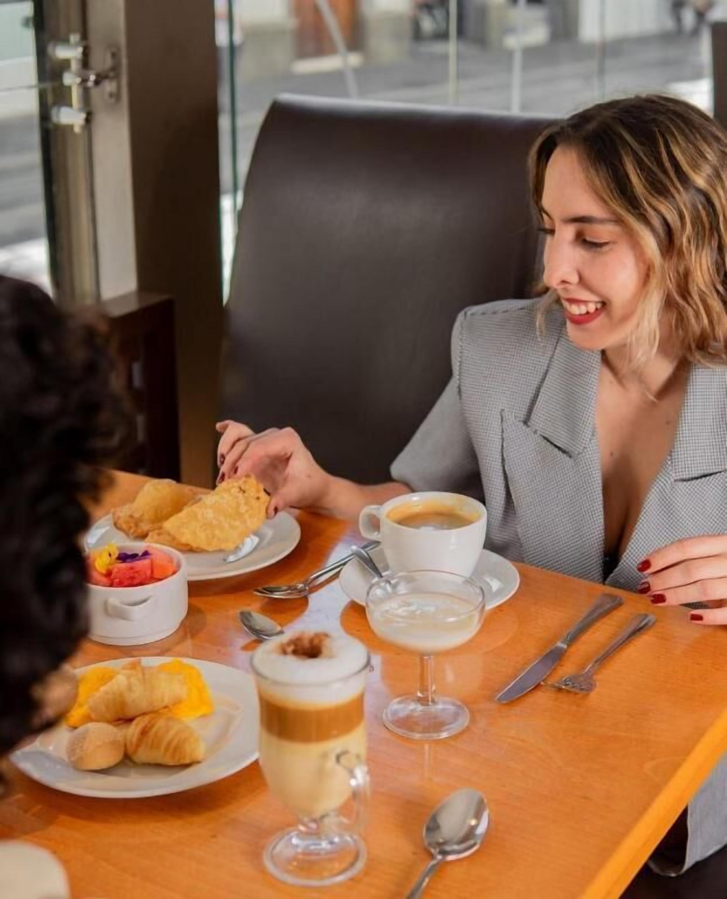 A person seated at a table with a meal consisting of croissants, a coffee cup, and a glass of a layered beverage.