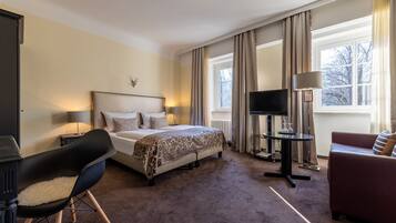 A hotel room with a bed, a TV, a desk, a chair, a sofa, and a window with curtains.