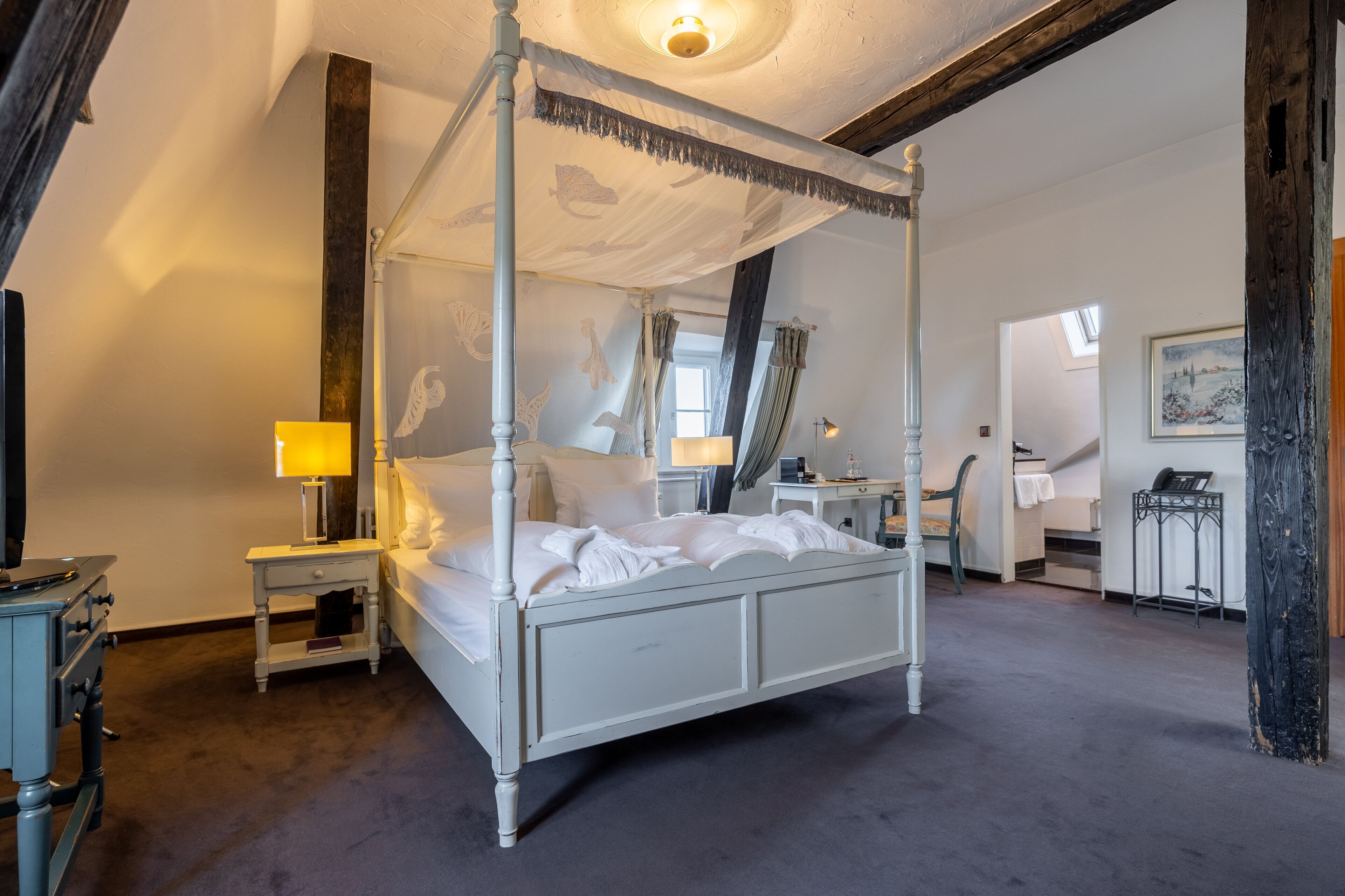 A four-poster canopy bed with white linens, a bedside table with a lamp, and a desk with a chair in the adjacent room.