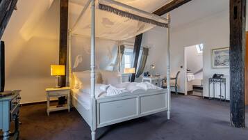 A four-poster canopy bed with white linens, a bedside table with a lamp, and a desk with a chair in the adjacent room.