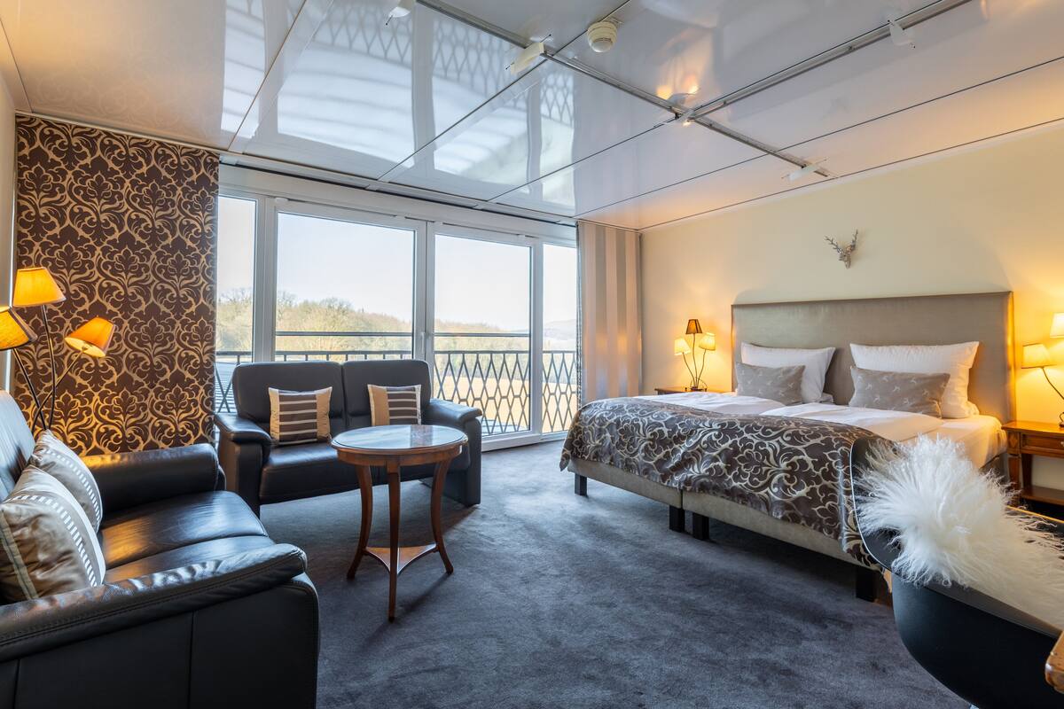A hotel room with a large bed, a sofa, a small table, and a view of the outdoors through large windows.