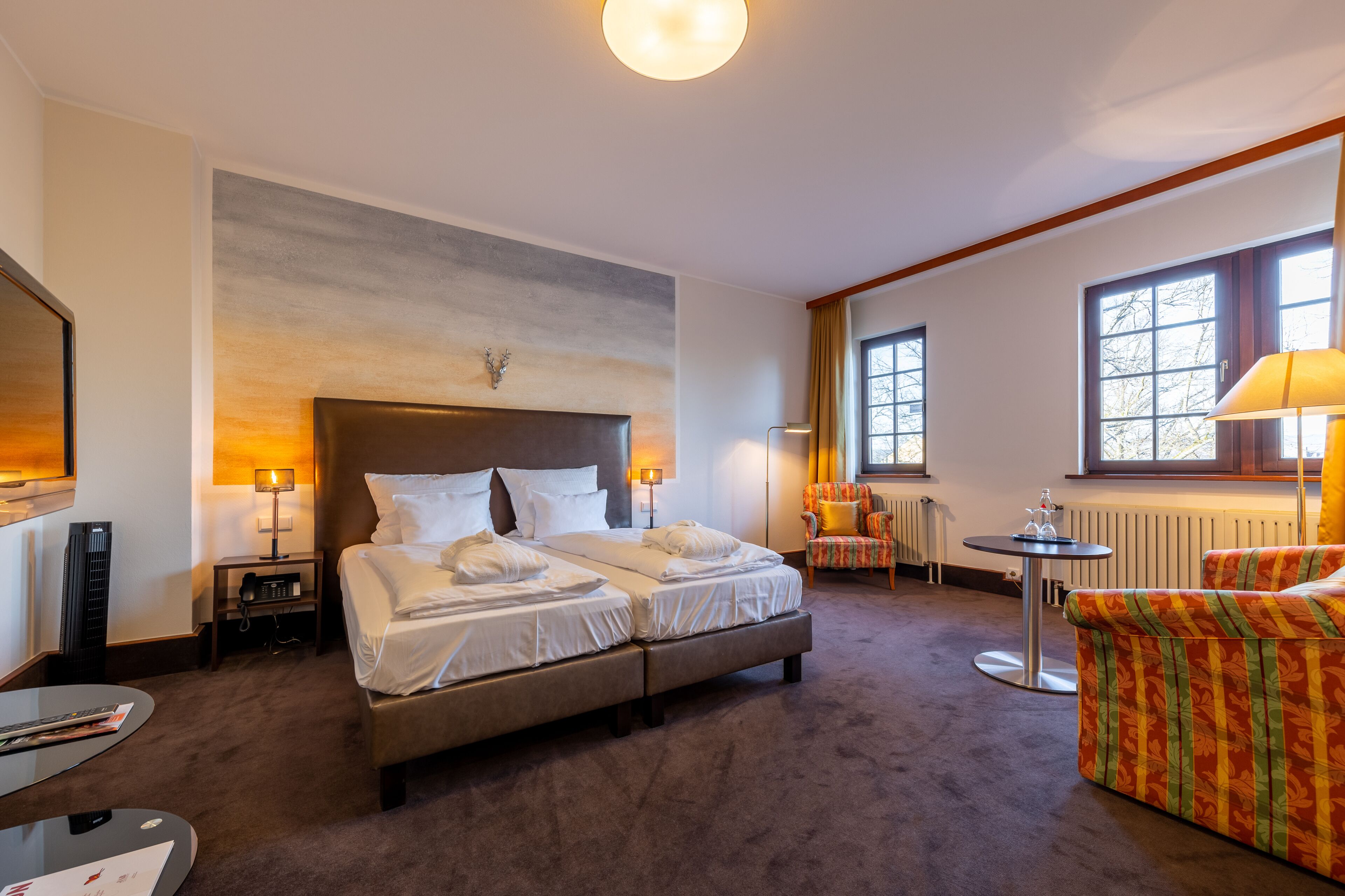 A hotel room with a large bed, two bedside tables, a chair, a small round table, and a radiator.