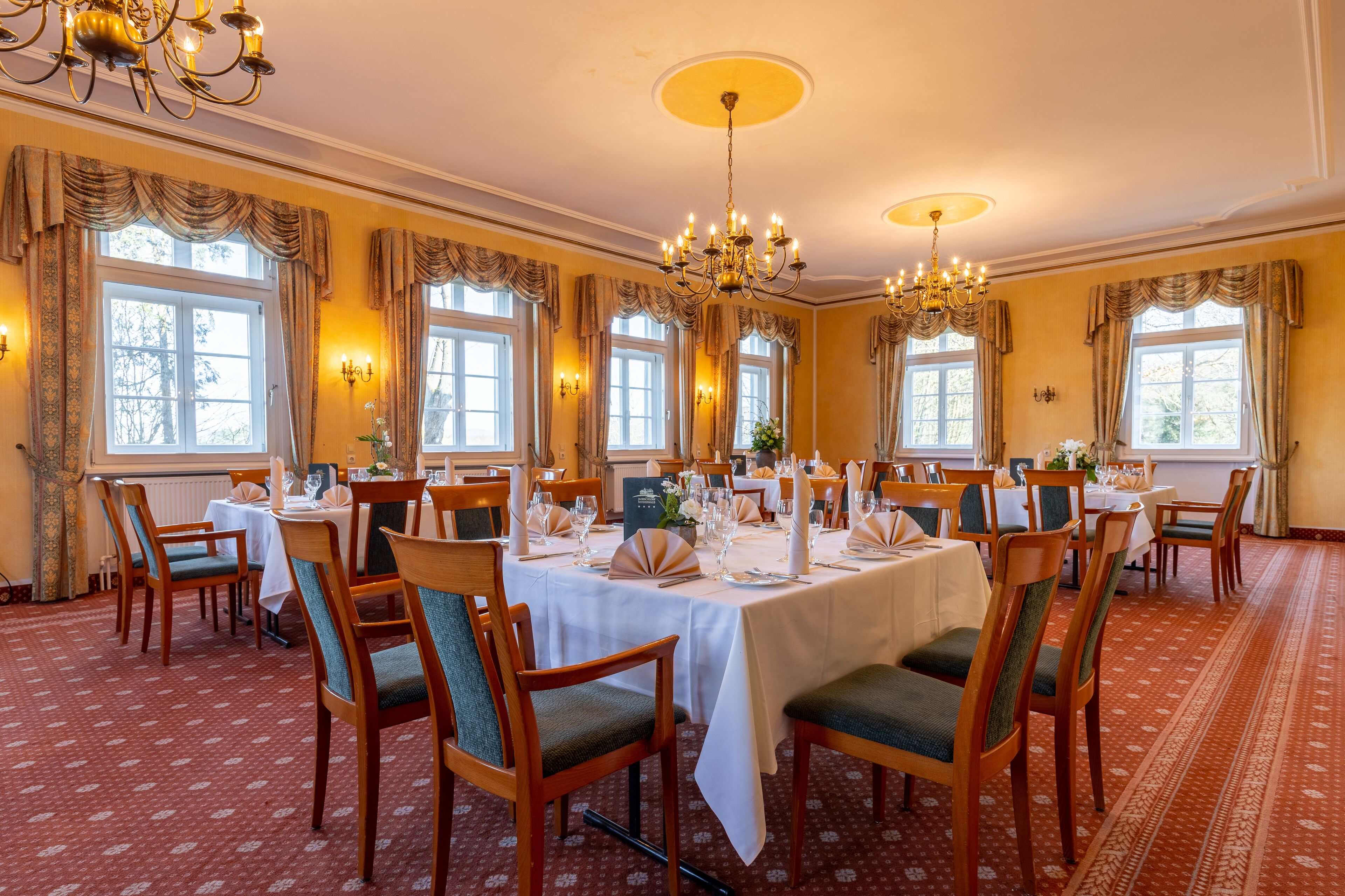 Un comedor formal con varias mesas dispuestas para el servicio, candelabros elegantes y ventanas grandes con cortinas.