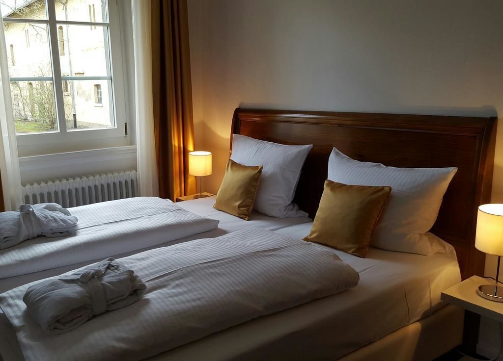 A hotel room with two beds, each with white linens and pillows, a wooden headboard, and a window with curtains.