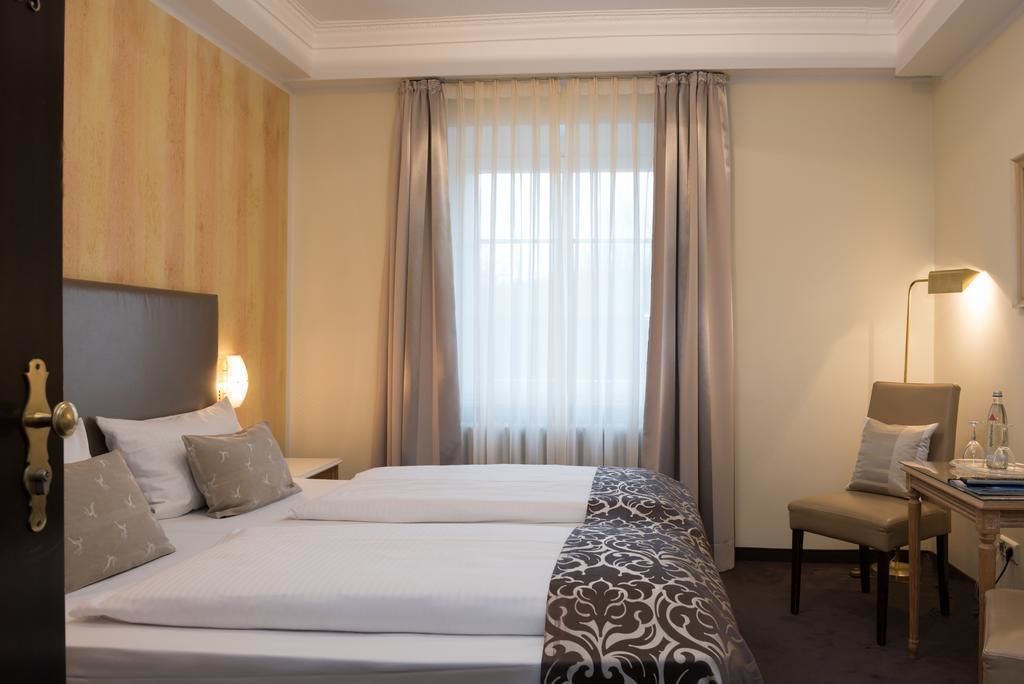 A hotel room with two beds, a desk with a chair, a lamp, and a window with curtains.