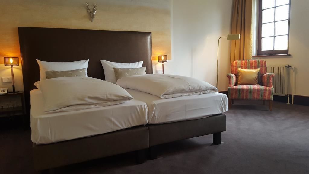 A double bed with white linens, two bedside lamps, a plaid armchair, and a window with curtains.