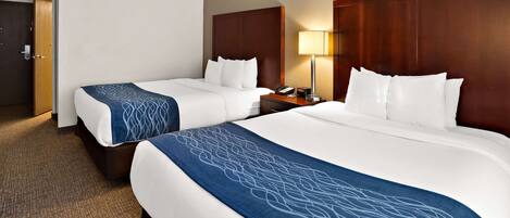 Habitación de hotel con dos camas, cada una con cabecero de madera, ropa de cama blanca y una manta estampada azul. Se observa una mesita de noche con una lámpara y un teléfono a un lado.