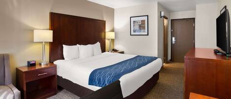 A hotel room with a large bed, a desk, a chair, a TV, and a lamp.