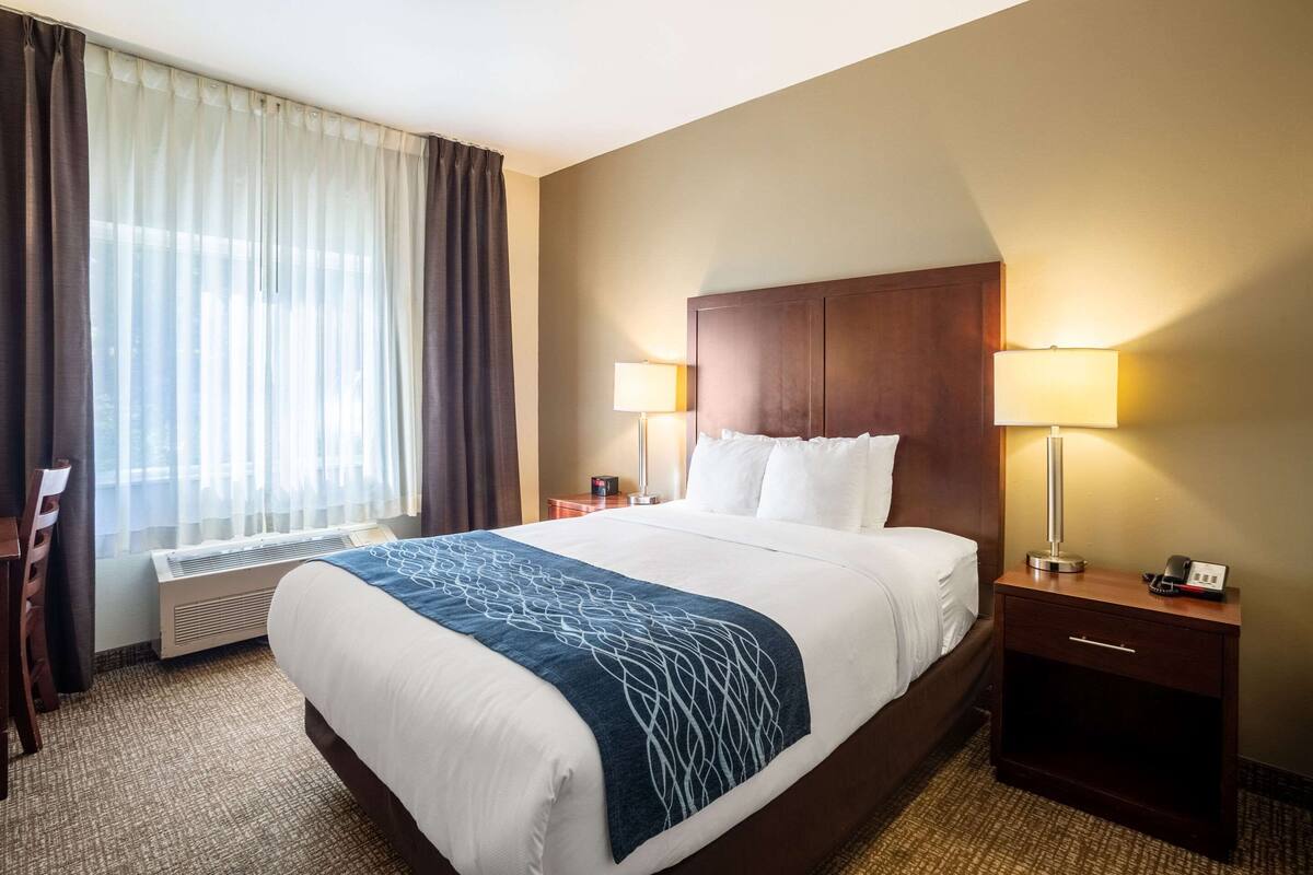 A hotel room with a large bed, two bedside tables with lamps, a chair, a heater, and a window with curtains.