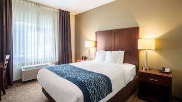 A hotel room with a large bed, two bedside tables with lamps, a chair, a heater, and a window with curtains.